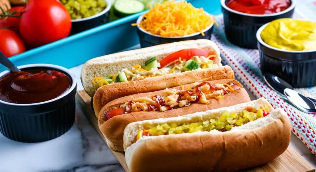 Three different fancy hot dogs on a tray with toppings in the background.