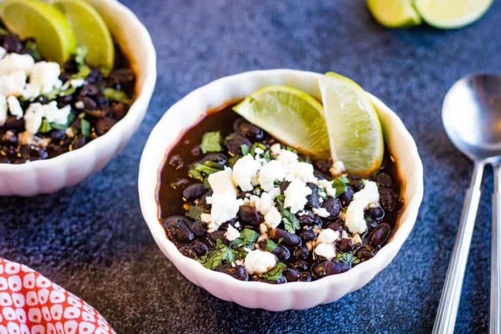 Low angle shot of two bowls of Mexican black beans garnished with crumbled cheese and lime wedges.