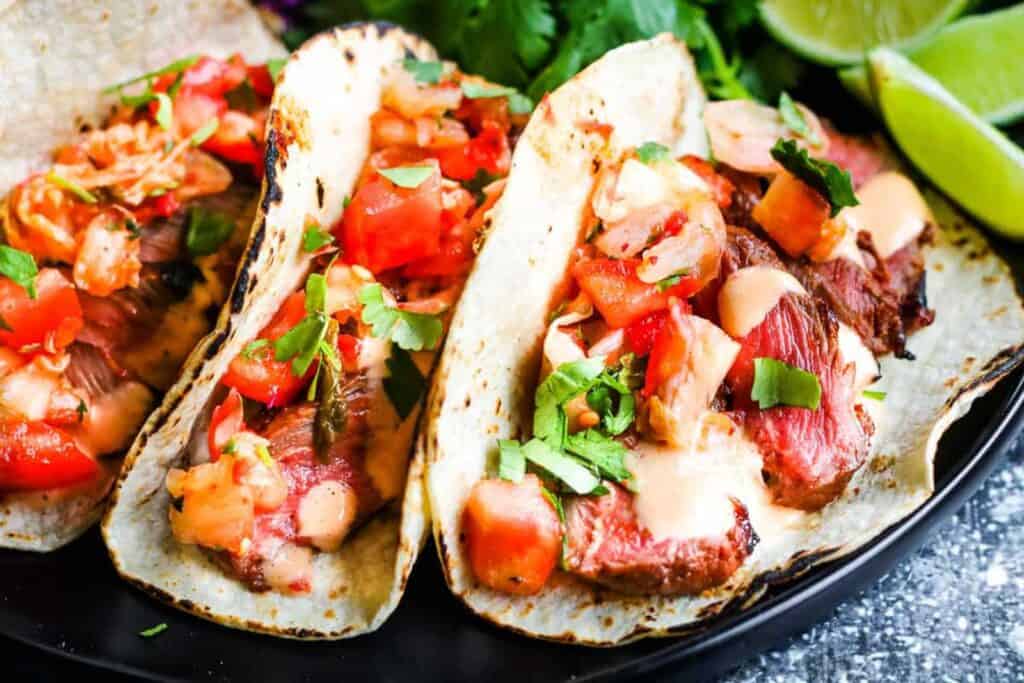 Horizontal shot of 3 korean tacos: corn tortillas filled with sliced steak, kimchi and pico de gallo salsa, and chopped cilantro. In the background are lime wedges, sliced red cabbage, and cilantro leaves.