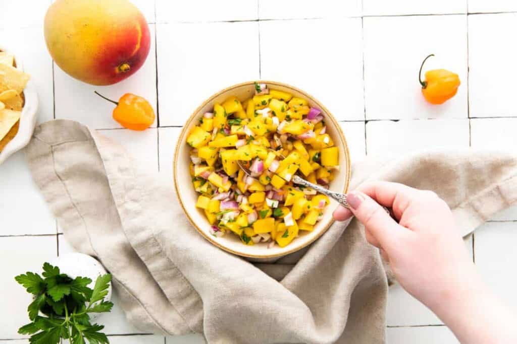 Overhead shot of a bowl of mango habanero salsa with a hand lifting a spoonful of the salsa.