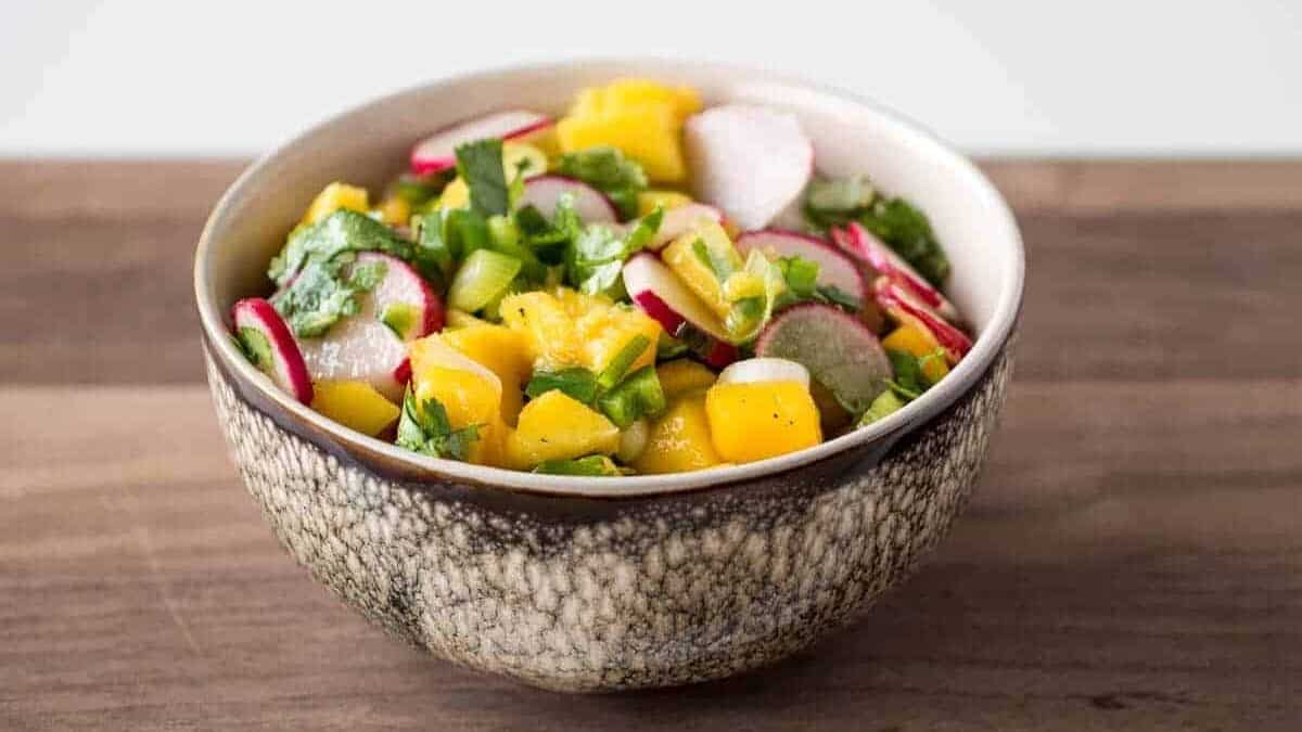 Radish mango salsa with cilantro and green onions in a brown and white bowl on a wooden cutting board.