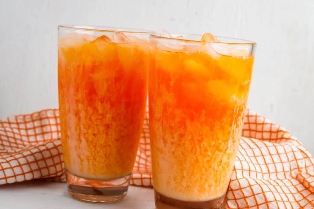 Two glasses of orange creamsicle soda with ice in front of an orange napkin.