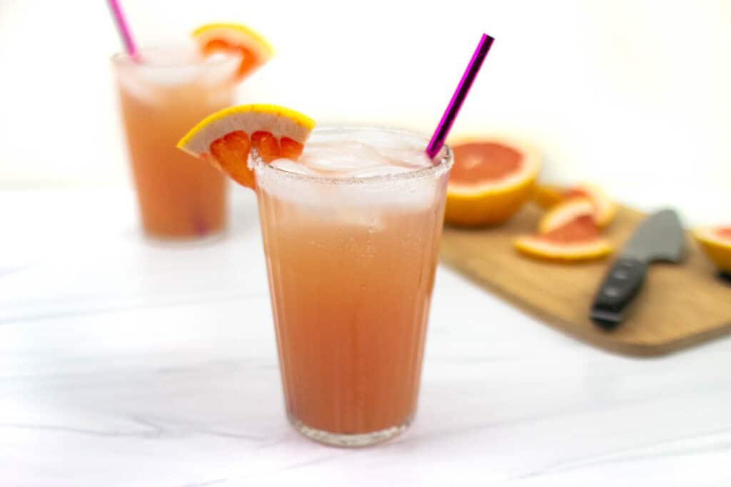 Two pink cocktails garnished with a wedge of grapefruit sit alongside a cut grapefruit on a cutting board with a knife.