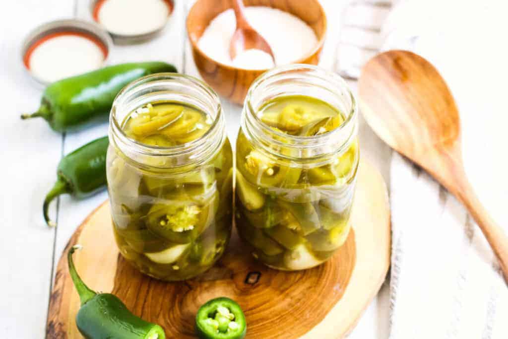 Two canning jars filled with pickled jalapenos on a wooden board with fresh jalapenos on the side.