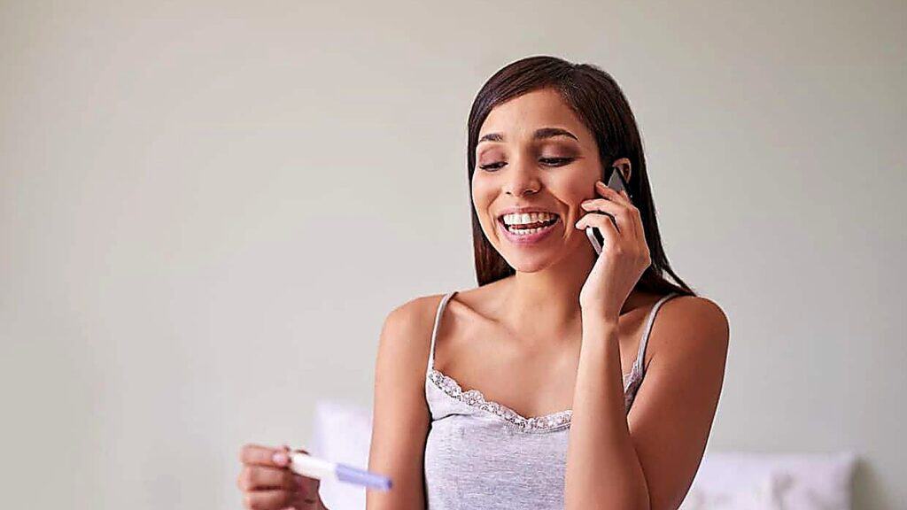 A woman smiles while talking on the phone and holding a pregnancy test, perhaps sharing her excitement as she discusses creative ideas on how to reveal pregnancy.