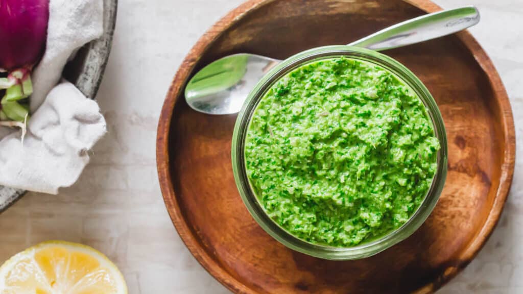 Radish greens pesto in a glass jar on a wooden plate with a serving spoon.