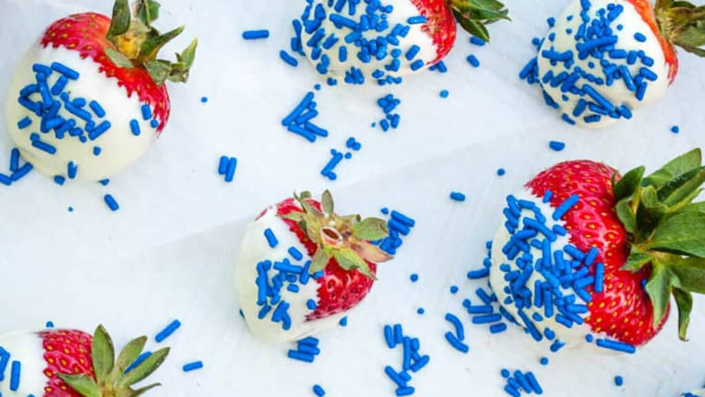 Red strawberrries coated with white chocolate with blue sprinkles on a baking sheet lined with parchment paper.