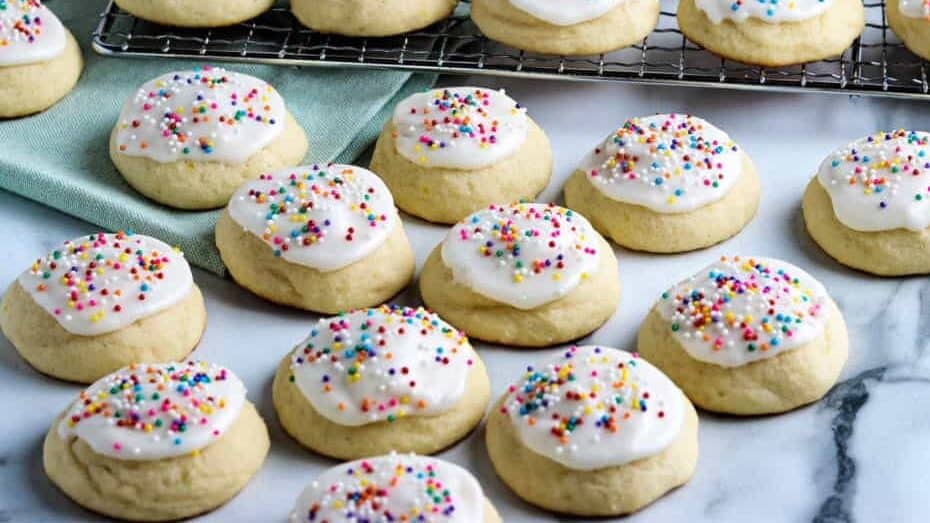 Ricotta cookies with icing and colored sprinkles on a cooling rack and on the counter.