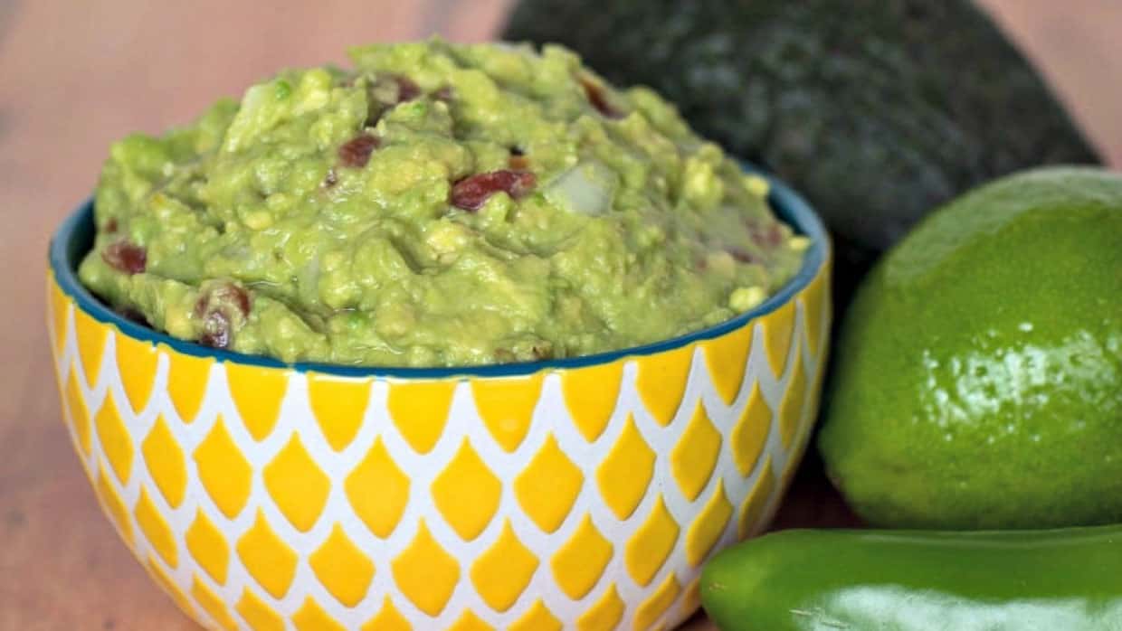 Yellow bowl with guacamole with avocado, and jalapenos next to it.