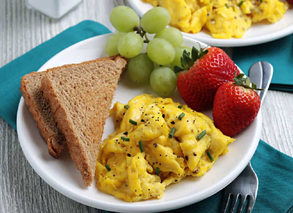 A white plate with scrambled eggs, toast and fruit.