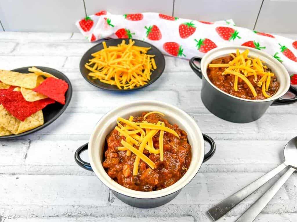 An image of Smoked Brisket Chili in two bowls with cheese and chips in the background.