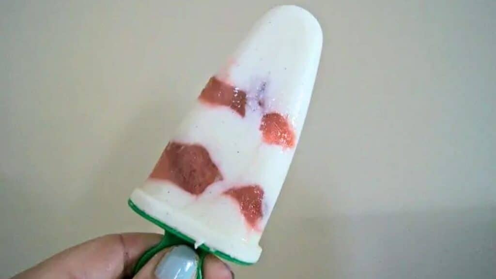 Image shows a hand holding a Greek yogurt popsicle against a plain background.