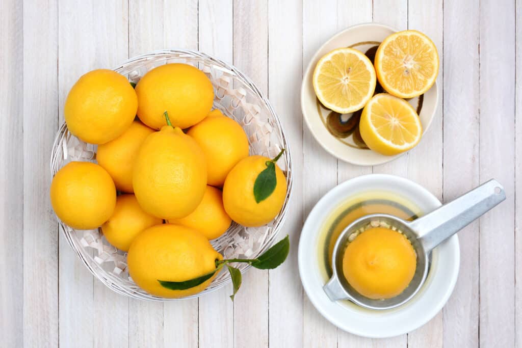 a glass bowl of lemons, a bowl of cut lemons and a citrus juicer with a lemon in it on a white wooden table.