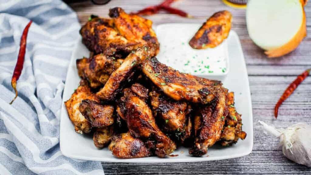 Pile of BBQ chicken wings on a white plate with dipping sauce.