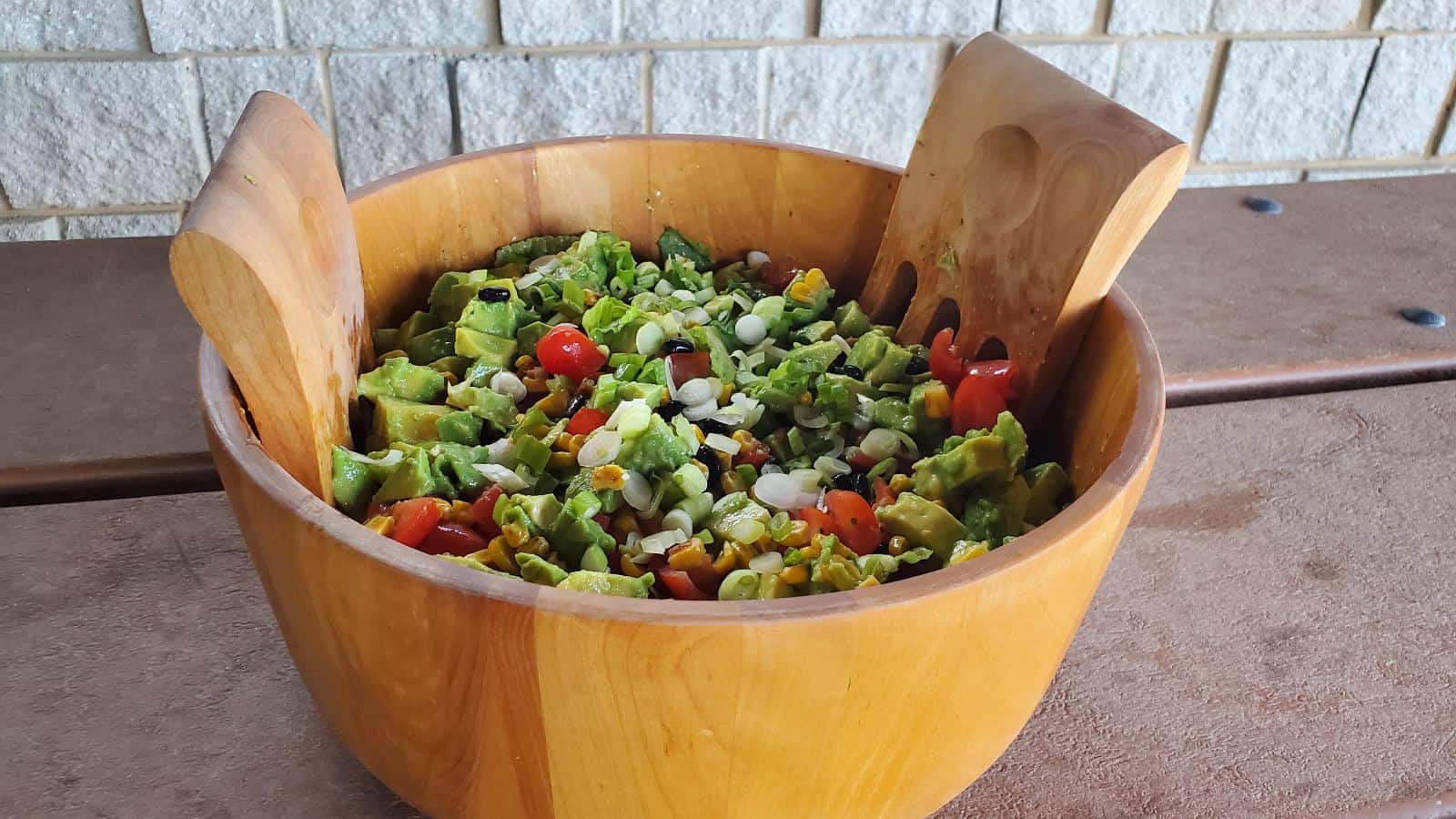 Image shows a wooden bowl containing avocado tomato salad shown from overhead and sitting on a picnnic table.