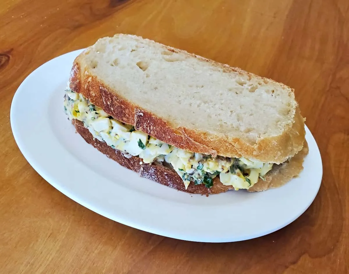 Image shows a bacon egg salad sandwich on a white plate sitting on a wooden table.