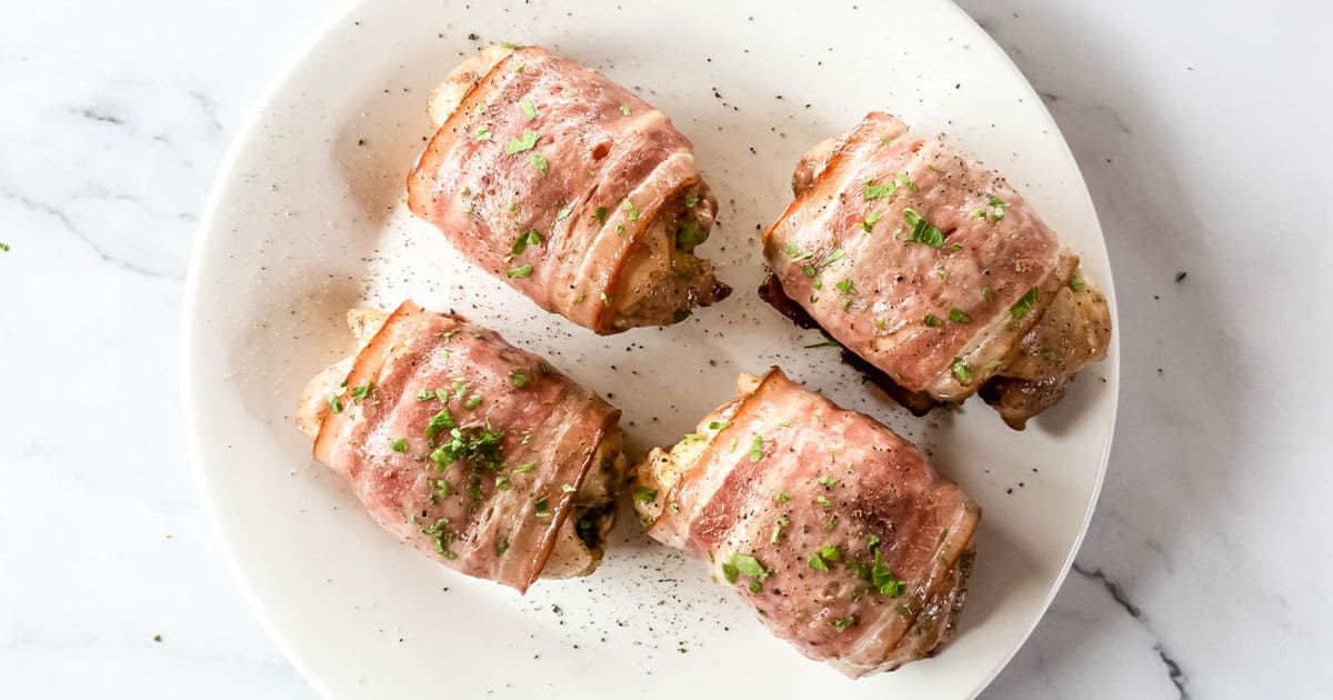 Four bacon-wrapped chicken thighs with parsley garnish on white plate.
