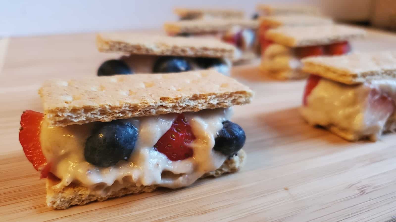 Image shows Banana ice cream sandwiches with strawberries and blueberries on a wooden board.