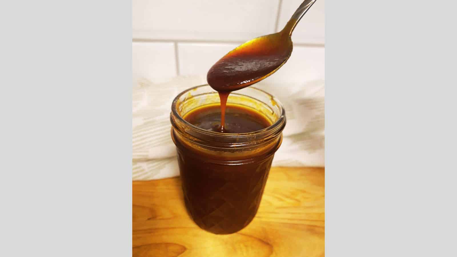 Barbecue sauce in clear jar with spoon dripping above.