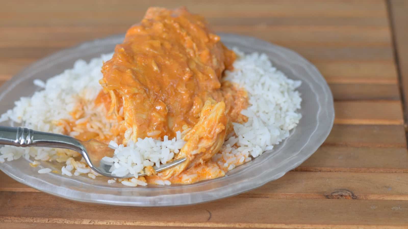 Image shows Bite of chicken paprikash on a fork sitting on a glass plate on a wooden table.