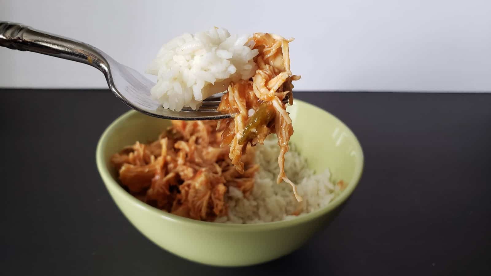 Image shows a Bite of tomato lime chicken on a fork with rice and the rest of the bowl in the background.