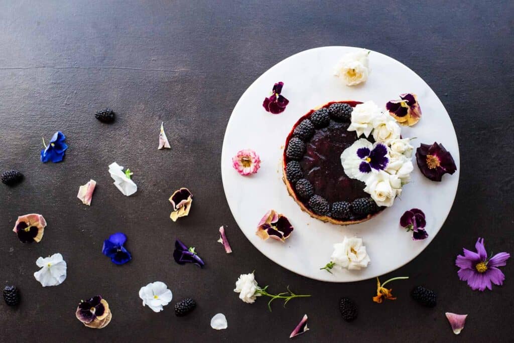 Overhead shot of a blackberry lime tart on a white plate surrounded by edible flowers. 
