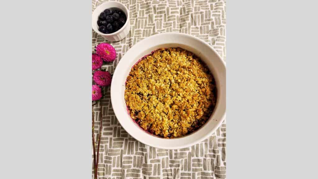 Blueberry crisp in round white dish next to pink flowers and bowl of blueberries.