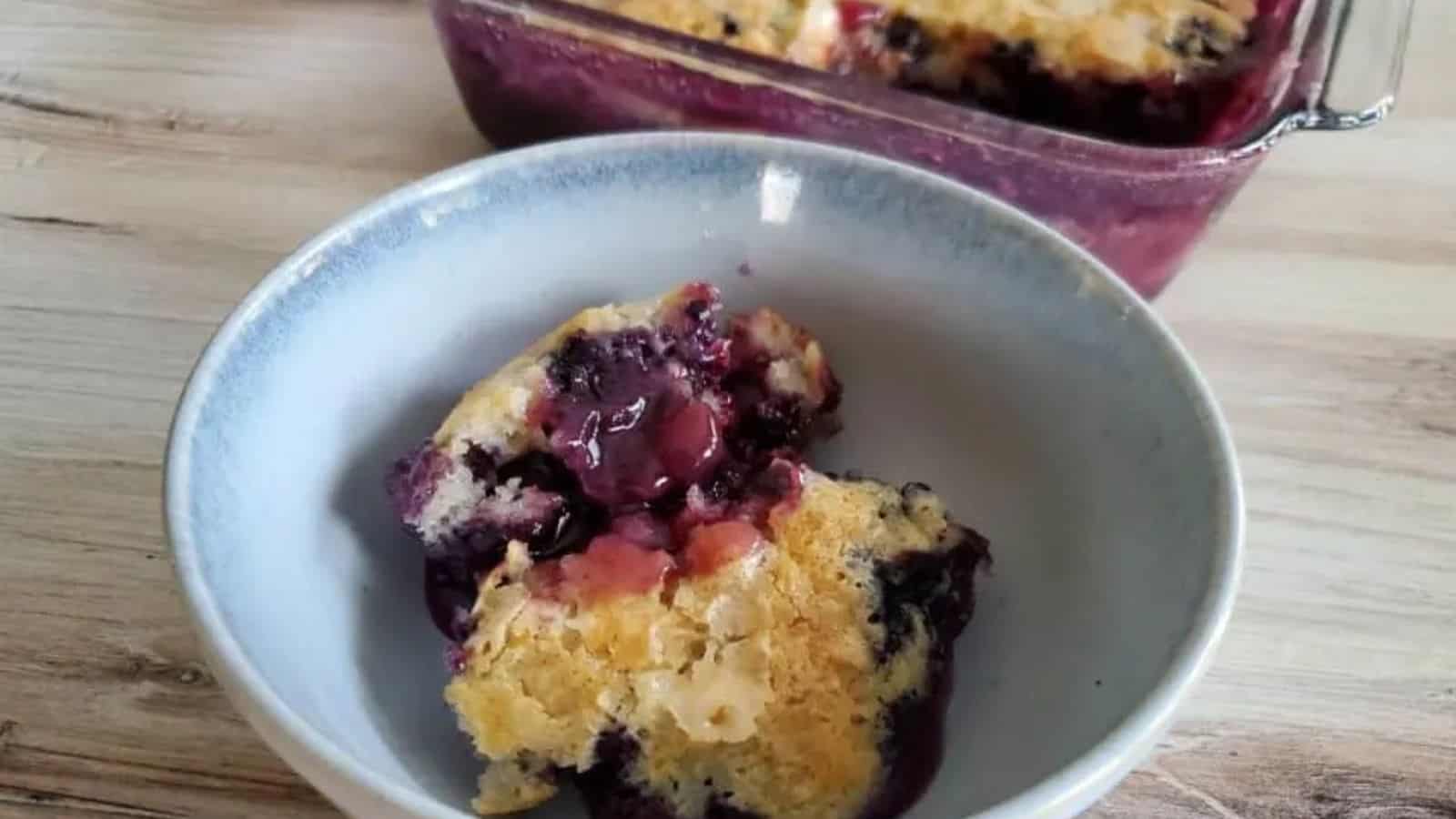 Image shows Blueberry Pudding Cake in a blue bowl with the full dish behind it.