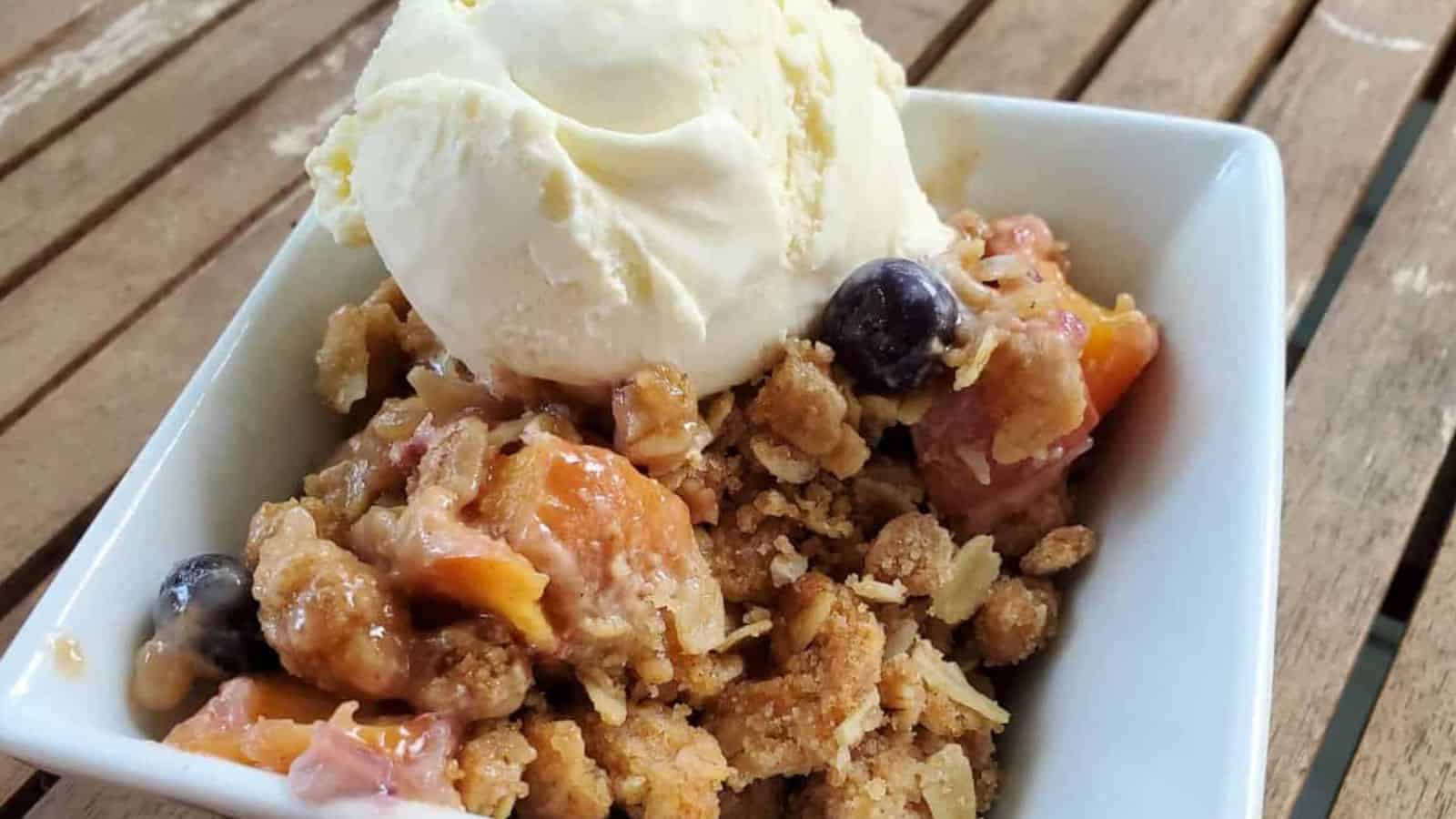 Image shows a white bowl with a serving of blueberry peach crisp topped with a scoop of ice cream and some whipped cream.