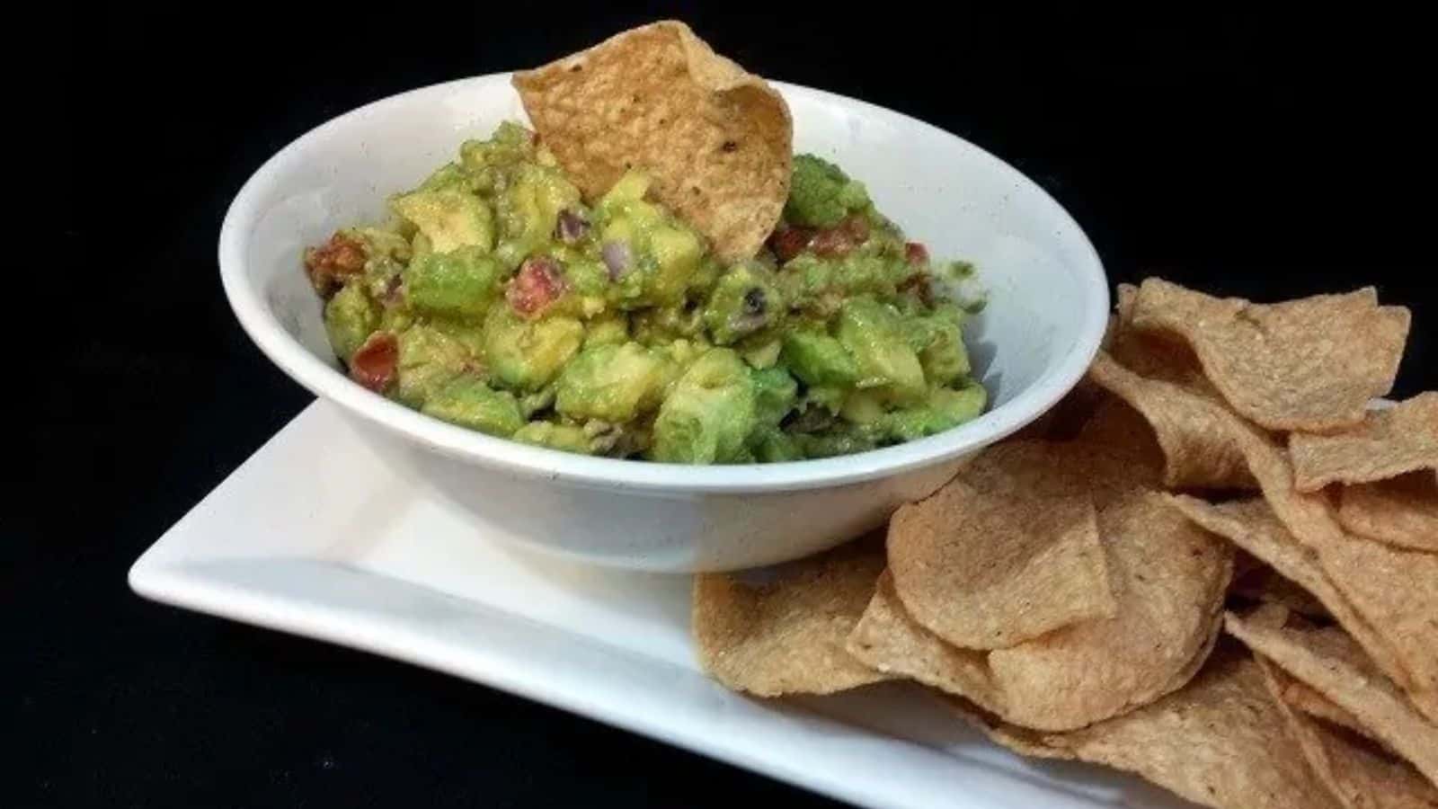 Image shows a white bowl with guacamole and a chip sticking in it with more chips on a tray to the right of it.