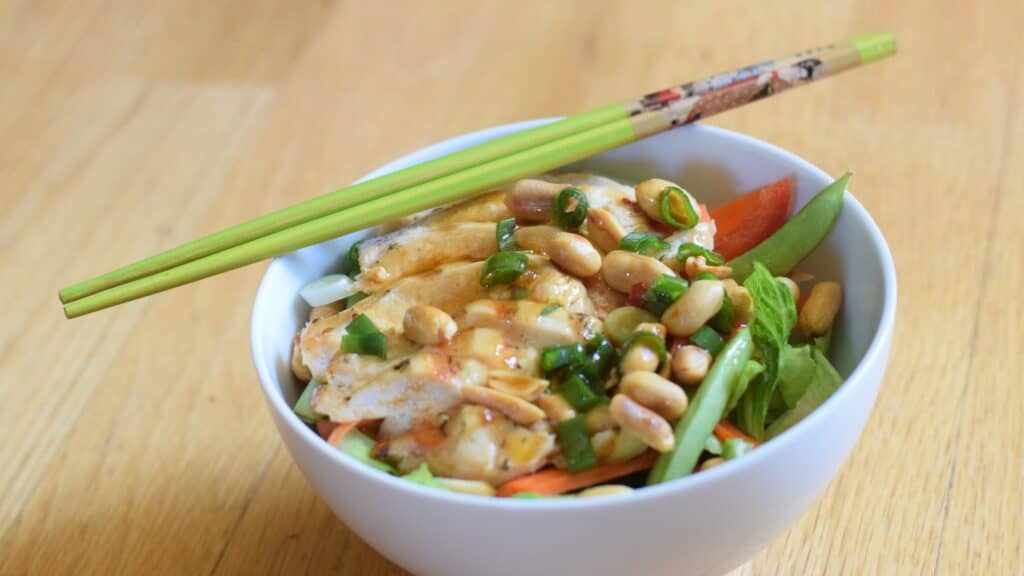 Image shows a Bowl of kung pao chicken salad with chopsticks on the edge.