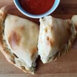 Image shows an overhead shot of a Calzone cut in half on a wooden board with a small bowl of marinara above it.