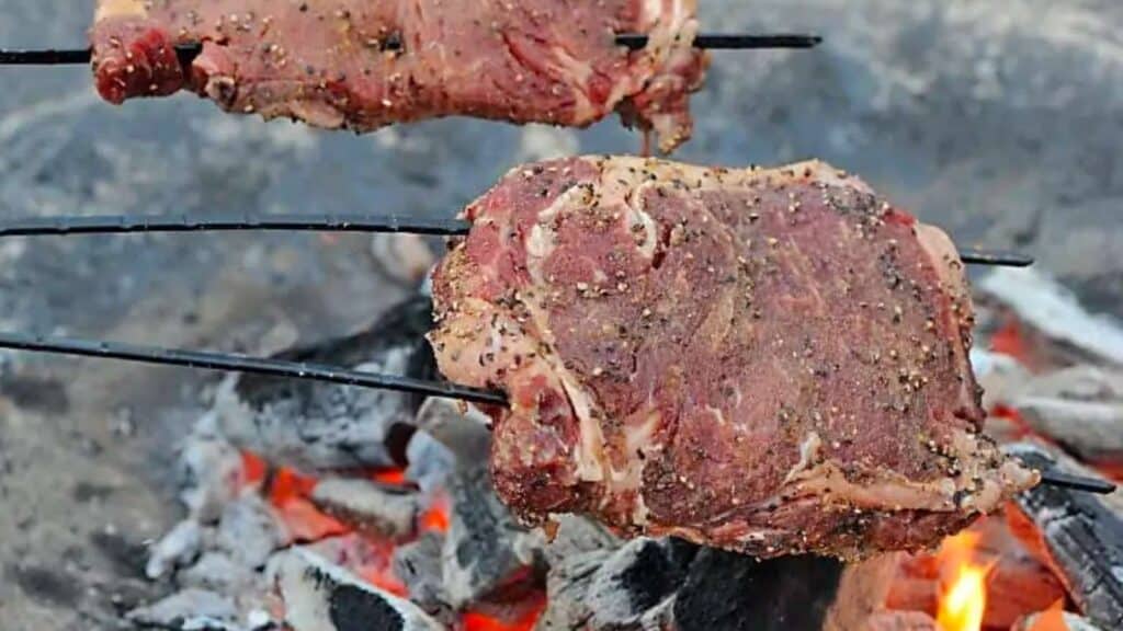 Two ribeye steaks on skewers over a campfire.