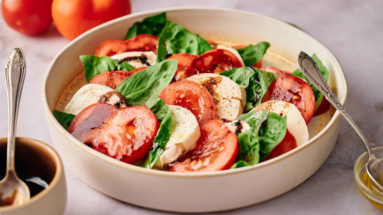 Caprese salad in a serving bowl, with tomatoes, oil and balsamic glaze nearby.