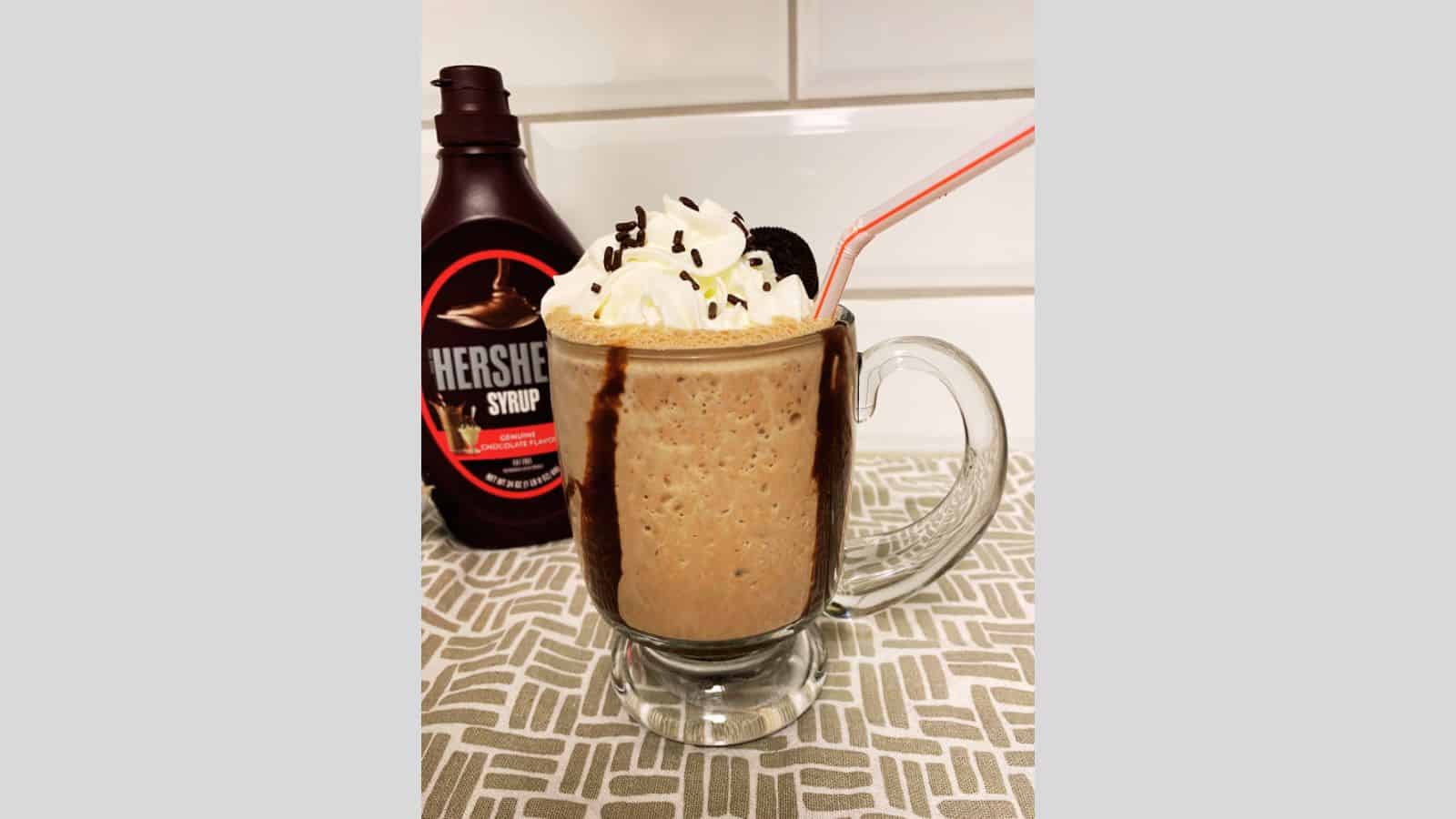 Chocolate milkshake in clear mug with whipped cream and Oreo cookie on top.