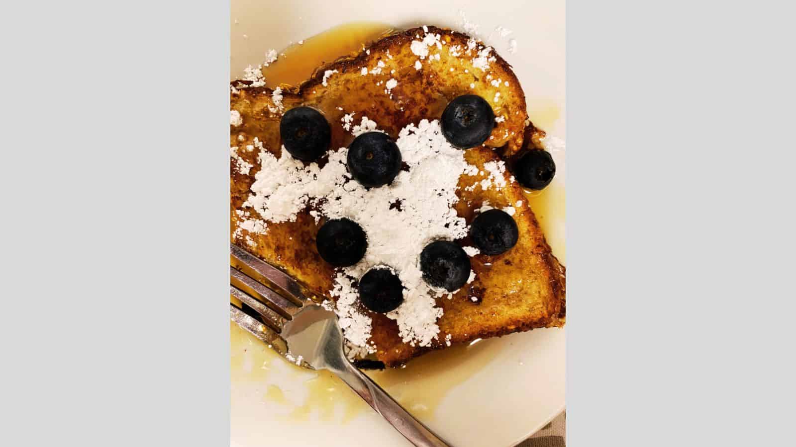 Slice of French toast on white plate topped with blueberries, powdered sugar, and syrup.