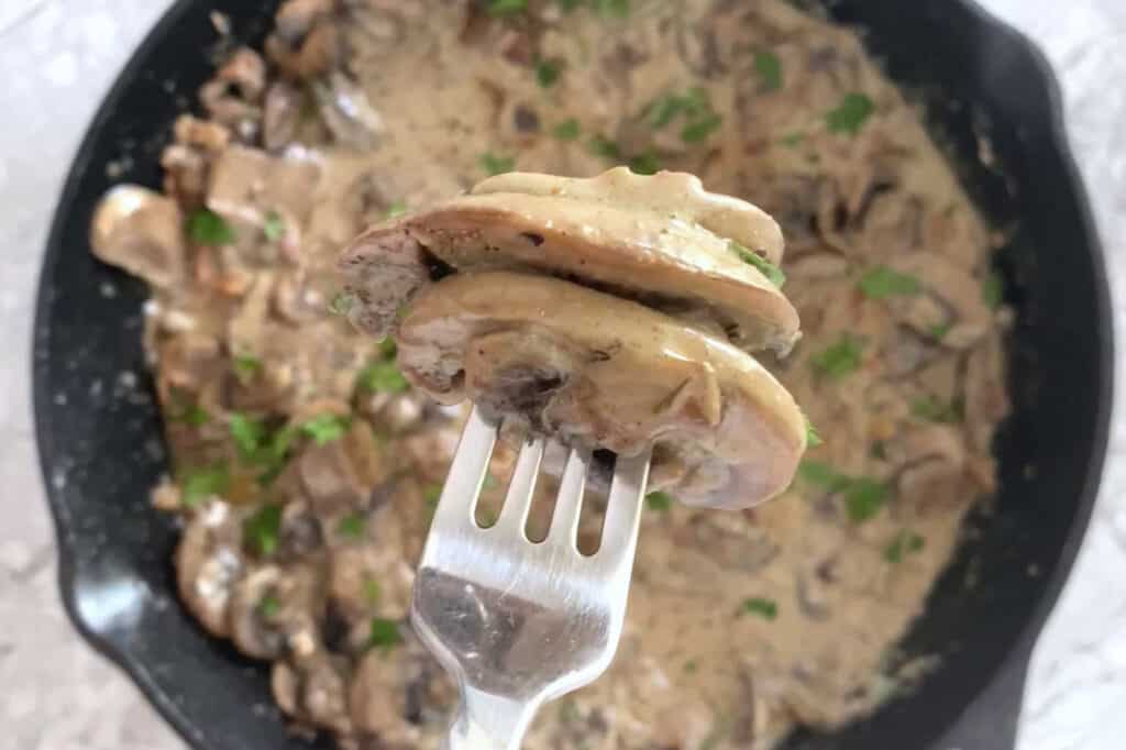 A fork with mushrooms held over a creamy mushrooms and bacon skillet.