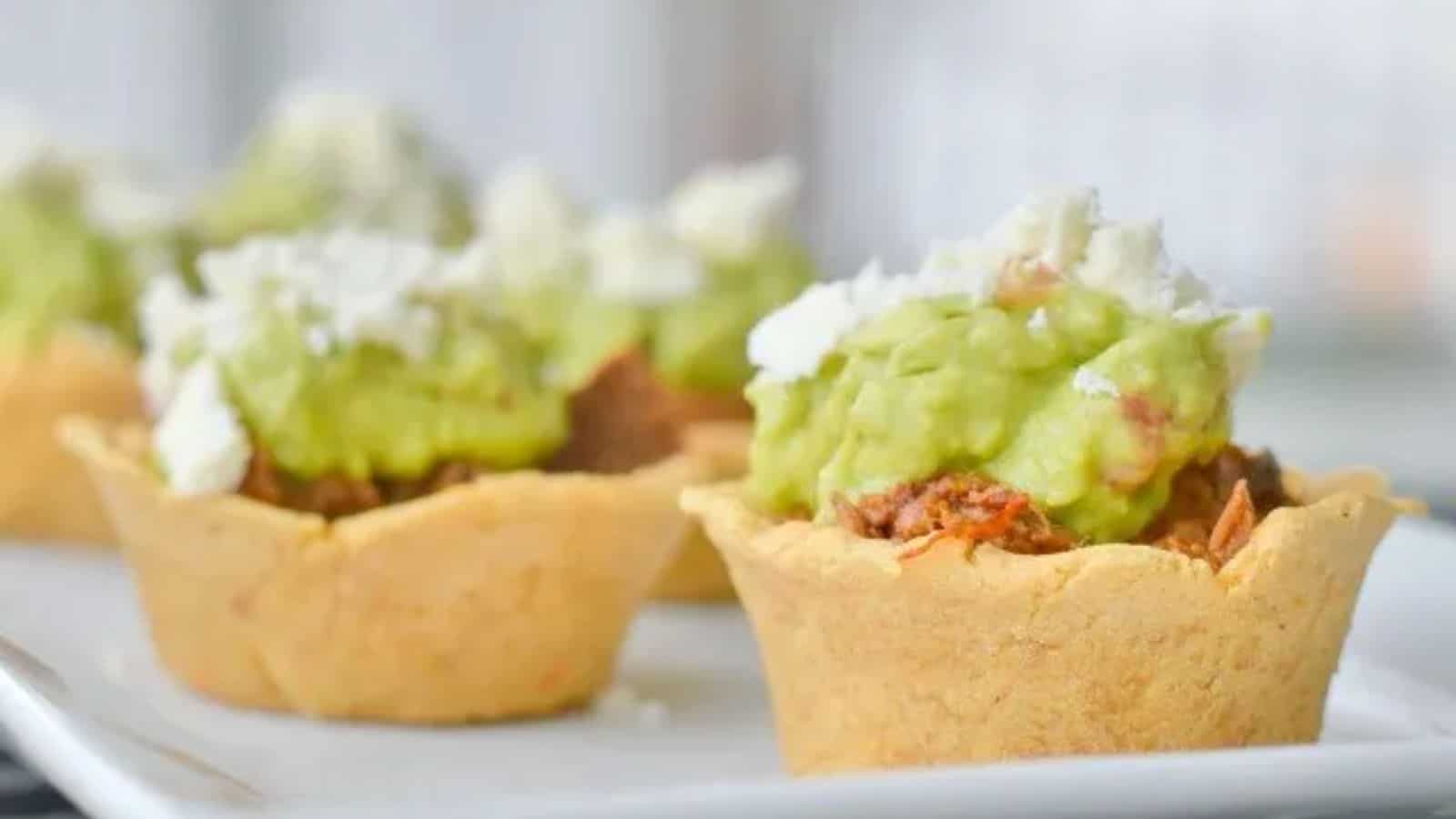 Image shows a close up head on view of Crockpot Mexican Beef in Masa Cups on a white tray, topped with guacamole and crumbled cotija cheese.