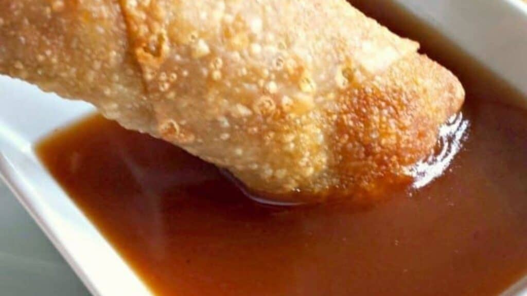 Image shows an Egg roll dipping into homemade sweet and sour sauce in a white bowl.