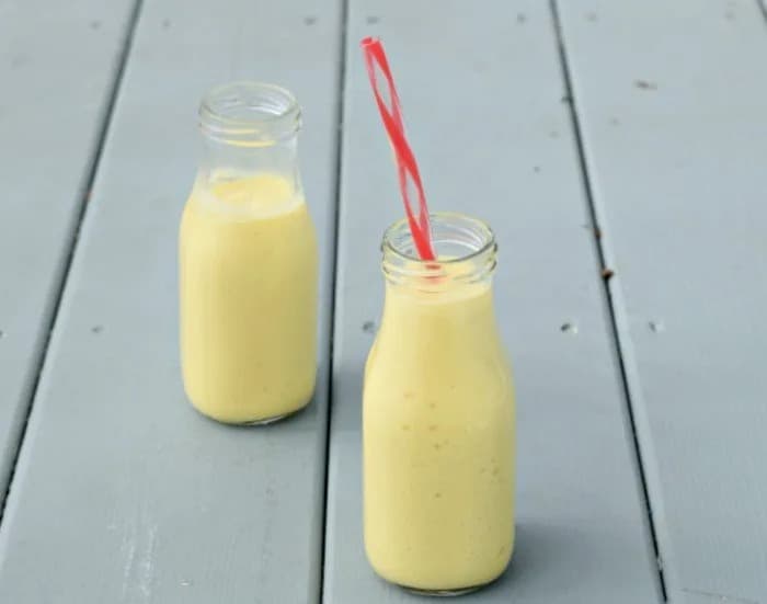 Two mango lassi drinks in glass milk bottles on a blue wood background with a red striped straw in the jar in the forefront.