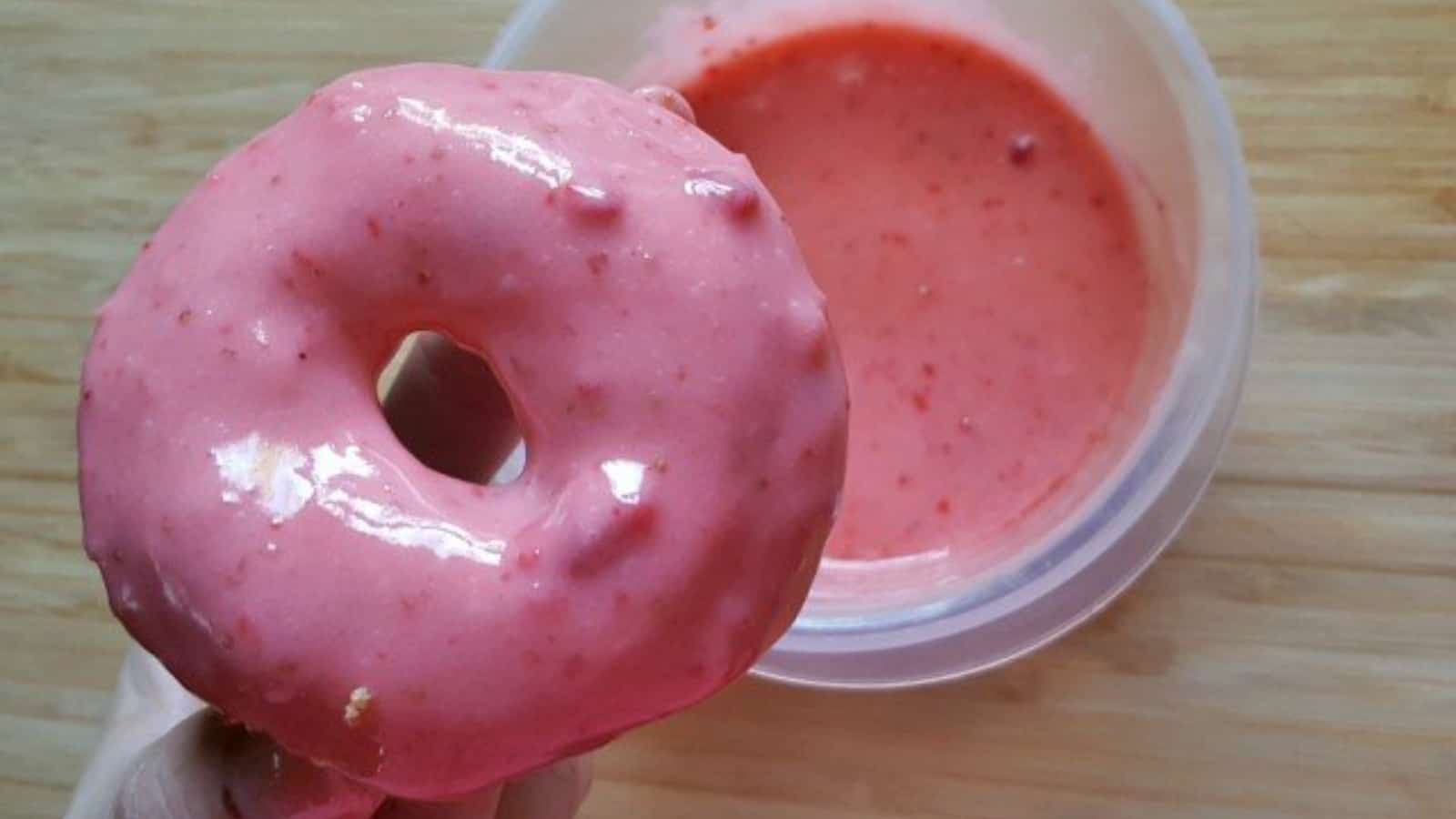 Image shows a freshly dipped strawberry donut held over the glaze.
