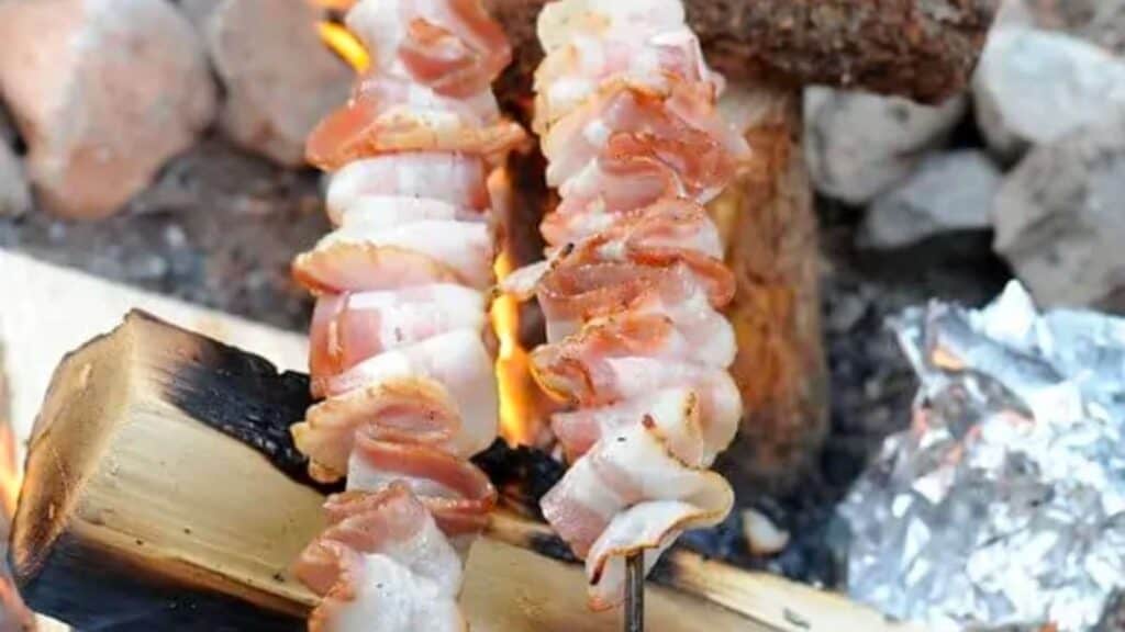 Bacon weaved onto two skewers over a campfire.