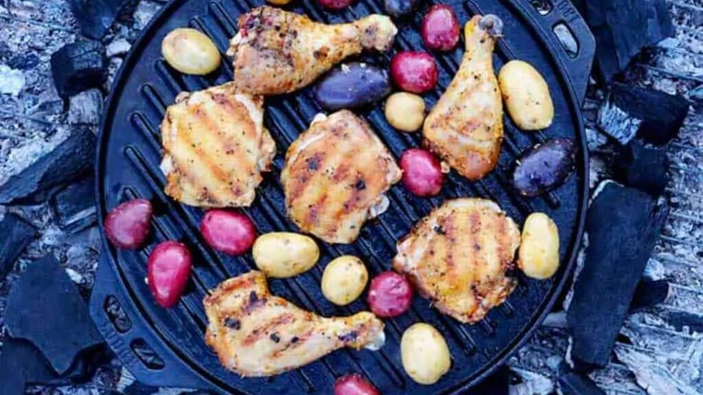 Grilled chicken thighs and legs on cast iron with rainbow potatoes over a campfire.