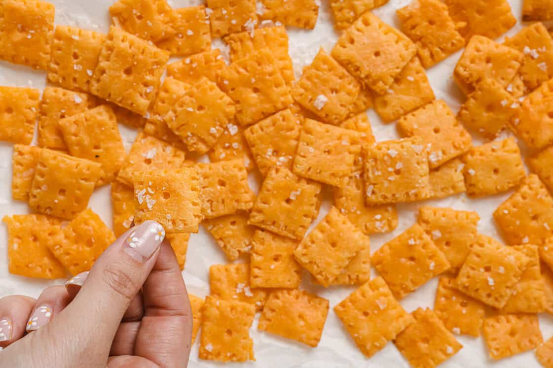 A hand holding a Cheez-It above a baking sheet of more crackers.
