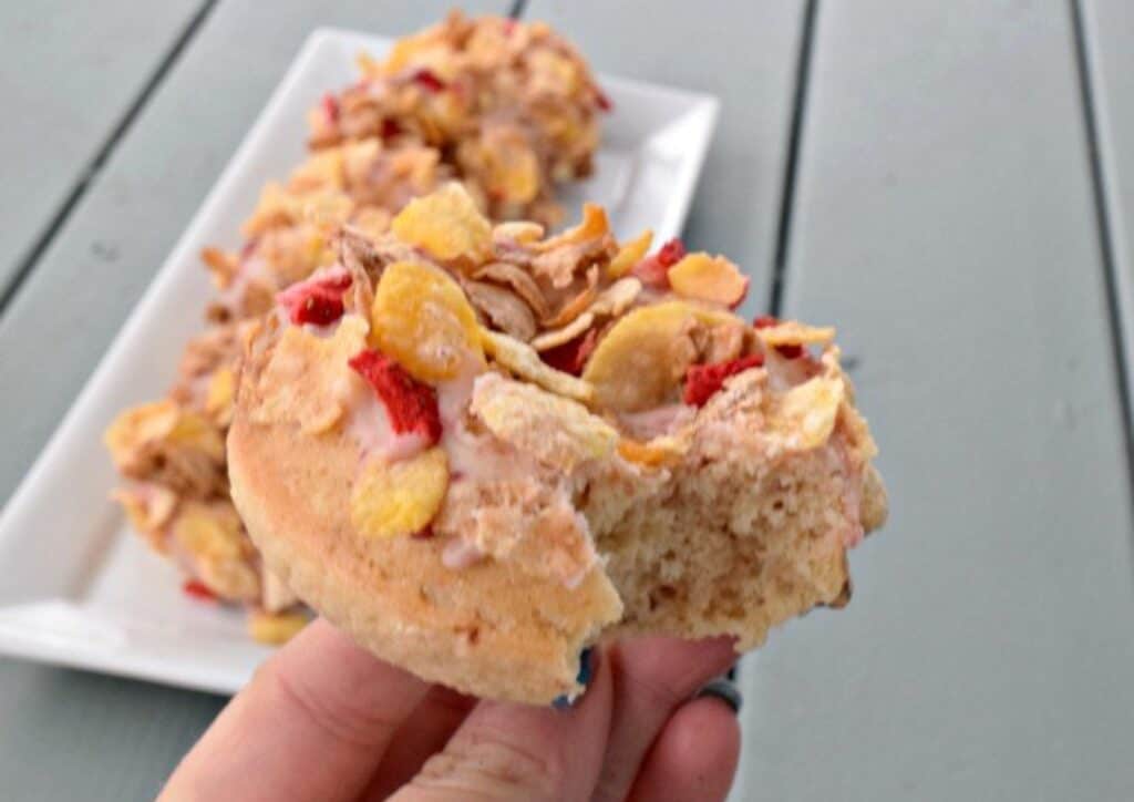 Image show a Hand holding a strawberry cereal donut with a bite taken out with more donuts on a white tray in the background.