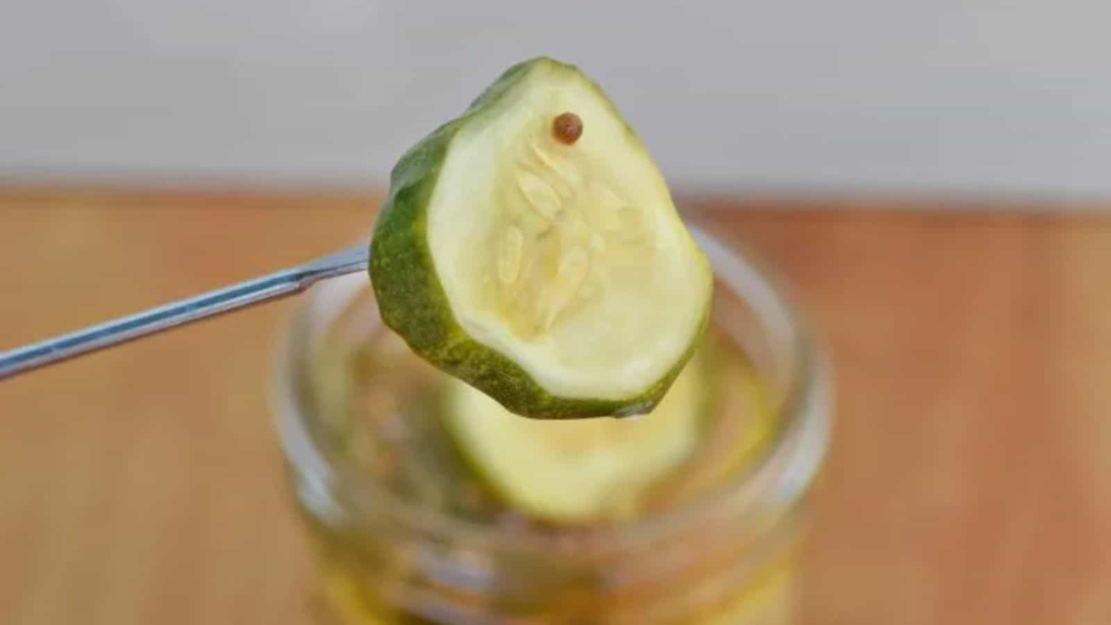 Image shows a skewer holding a bread and butter pickle with the full mason jar behind it.