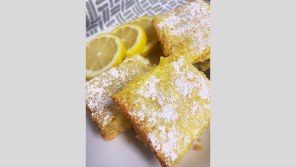 Close up of lemon bars on white plate with sliced lemons in background.