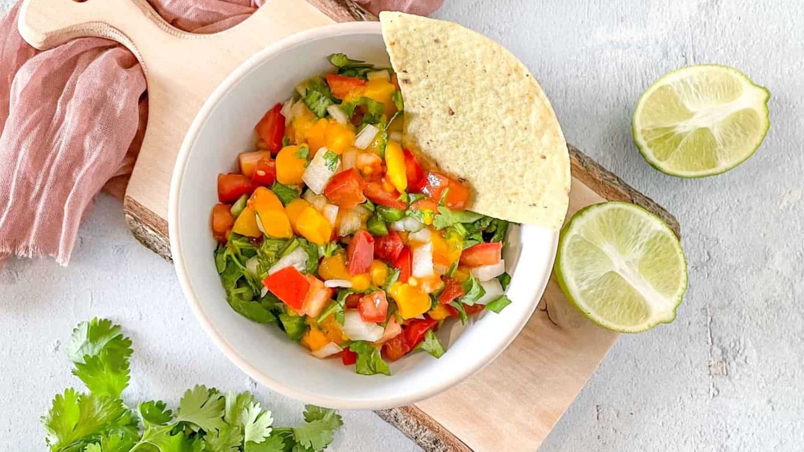 A bowl of mango pico de gallo sits on a rustic wooden cutting board surrounded by limes, cilantro, and a pink linen.