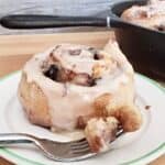 Image shows a cinnamon roll on a plate with a fork holding a bite and the full pan behind it.