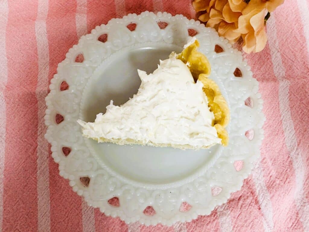 Slice of coconut cream pie on white plate with pink tablecloth in background.